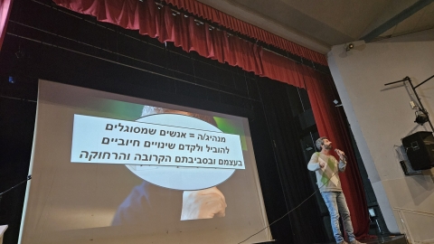 איש עומד על במה ומחזיק מקרופון ברקע מוקרנת מצגת.jpg