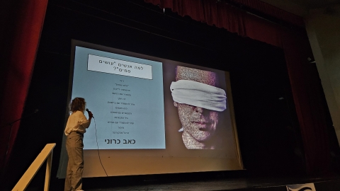 אישה עומדת על במה ומחזיקה מקרופון ברקע מוקרנת מצגת.jpg