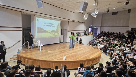 שתי תלמידות עומדות על במה ומדברות למיקרופון. סביבן יושבים ילדים.jpg