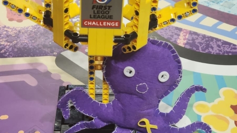 גביע שעשוי מחלקי לגו ועליו כתוב  FIRST LEGO League challenge לידו בובת תמנון בצבע סגול ועליה סיכת הח