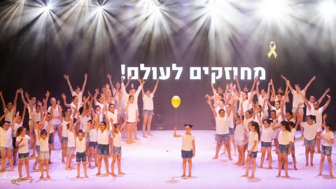 ילדים עומדים על הבמה לובשים חולצות לבנות. ילדה אחת מחזיקה בלון בצבע צהוב. על מסך מאחוריה מופיע המשפט