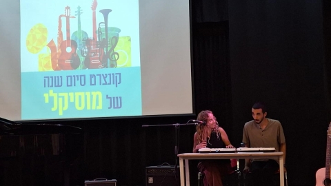 אישה ואיש מנגנים על במה. מאחוריהם מצגת שכתוב עליה קונצרט סיום השנה של מוסיקלי.jpg