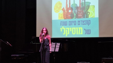 אישה שרה על במה. מאחוריהם מצגת שכתוב עליה קונצרט סיום השנה של מוסיקלי.jpg