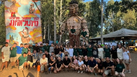 תמונה של ילדים מאחוריהם מייצג מסנדות ולידם פוסטר ענק עם דמויות מצויירות עליו וכתוב כפר שמואל מציגים 