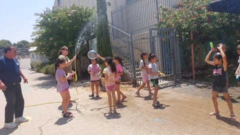 ילדים משחקים במים ומורה מחזיקה צינור מים ומשפריצה עליהם.jpg