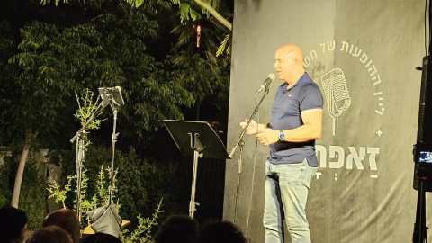 איש בחולצה כחולה עומד על הבמה ומחזיקה מיקרופון.jpg