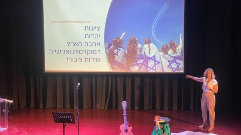 אישה עומדת ומחזיקה מיקרופון ומאחוריה מצגת שכתוב עליה ציונות יהדות אהבת הארץ דמוקרטיה ואנושיות שירות 