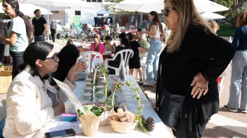 אישה יושבת לידה שולחן ועליו עציצים. אישה אחרת עומדת מולה.jpg