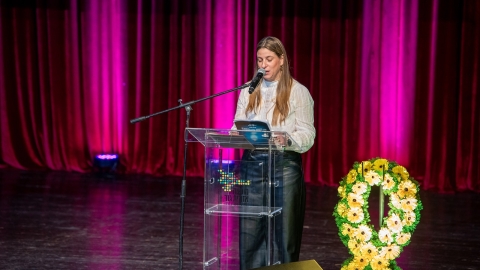 ישה לובשת חולצה לבנה. עומדת על במה ונואמת. לידה סמל החטופים מפרחים צהובים.jpg