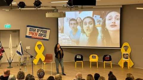 מירב לשם גונן עומדת על הבמה ומספרת סיפורה האישי. מאחוריה כיסאות צהובים וכתוביות להחזיר את כולם הביתה