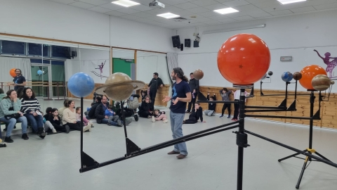 ילדים והורים יושבים בתוך אולם ומקשיבים למרצה ומסתכלים על מודל של מערכת השמש.jpg