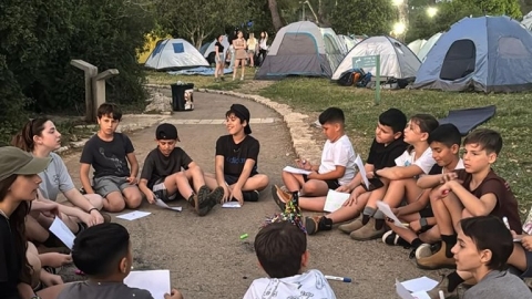 קבוצת חניכים יושבת במעגל בשח המחנה ומאחוריה אוהלים.jpg